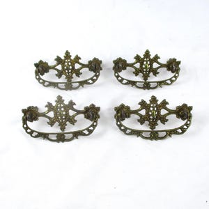 May include: Four matching antique brass drawer pulls featuring an ornate, openwork design with a central diamond-shaped motif and a curved, swinging handle. Each piece displays a dark, aged metal finish with intricate scrollwork and filigree details.