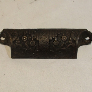 May include: A dark brown metal drawer pull with an ornate floral design. The pull has two holes for mounting.