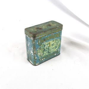 May include: Vintage rectangular tin box with a faded blue and green color scheme. The box features the word "CHAMPAGNE" in gold lettering, along with an illustration of a champagne bottle and grapes. The tin shows signs of wear and tear.