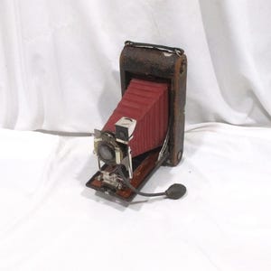 May include: Vintage brown leather camera with a red bellows. The camera has a silver lens and a black bulb shutter release. The camera is open and ready to use, showcasing its classic design and craftsmanship.