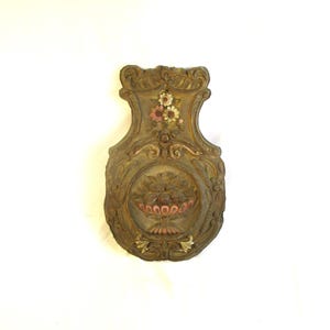 May include: Ornate, antique wall plaque with a carved design. The plaque features a central oval with a basket of fruit and flowers, surrounded by detailed scrollwork and floral accents. The color palette includes gold, brown, and touches of pink and white.