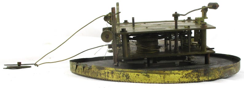 Peut inclure: Un mouvement d'horloge ancien avec des engrenages, des leviers et une base circulaire en laiton. Le m&eacute;canisme est expos&eacute;, r&eacute;v&eacute;lant les composants internes complexes. La base pr&eacute;sente un aspect us&eacute;.