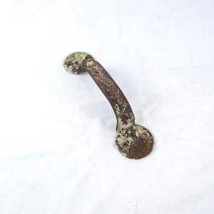 May include: A weathered metal door handle featuring a curved grip and two circular mounting plates at each end. The surface displays significant rust and remnants of chipped, off-white paint, showing a textured, aged appearance throughout.