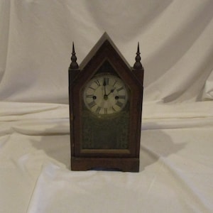 May include: A vintage wooden clock with a steeple-shaped design. The clock face has Roman numerals and a white background. The clock is in a dark wood frame.