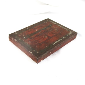 May include: A rectangular, antique metal box with a red and brown distressed finish. The box has text on the top, which is partially visible. The box is likely vintage and shows signs of wear.