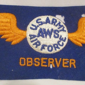 May include: A blue felt patch with orange embroidered wings and a white circle with the text "U.S. ARMY AIR FORCE" in blue. The text "OBSERVER" is embroidered in orange below the circle.
