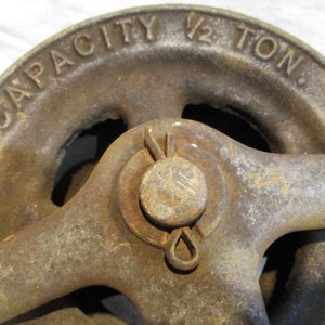 Pu&ograve; includere: Una carrucola in ghisa vintage con una manovella. La carrucola &egrave; etichettata "CAPACITY 1/2 TON."