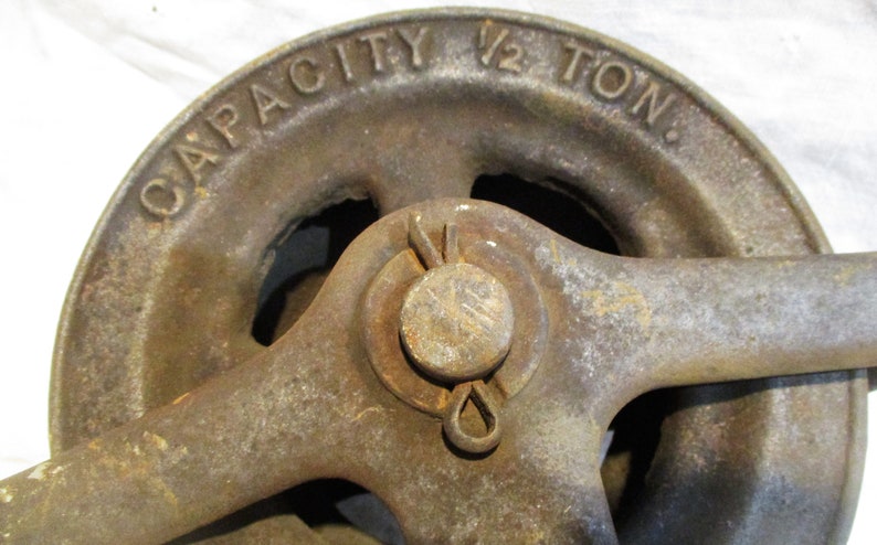 Pu&ograve; includere: Una carrucola in ghisa vintage con una manovella. La carrucola &egrave; etichettata "CAPACITY 1/2 TON."