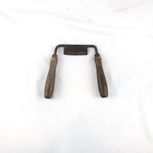 May include: A vintage drawknife featuring a central metal blade with a dark, weathered patina and two parallel wooden handles. The handles are cylindrical, showing signs of wear and age, connected to the blade by curved metal arms.