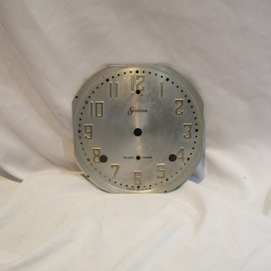 May include: A silver clock face with black hands and gold numbers. The clock face is labeled "Sessions" and has the words "Silent" and "Chime" printed below the 6. The clock face is made in the USA.