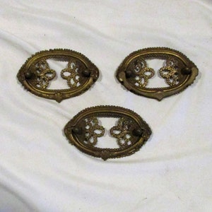 May include: Three antique brass drawer pulls with an ornate design. The pulls are oval shaped with a cutout design in the center.