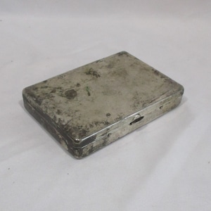 May include: A silver metal cigarette case with a worn, aged appearance. The case has a hinged lid and a small latch.