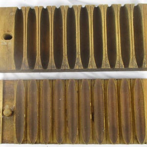 May include: A vintage wooden cigar mold consisting of two rectangular blocks with ten carved, elongated, concave channels. The top piece features a circular hole on the left side, while the bottom piece includes a small wooden peg for alignment.