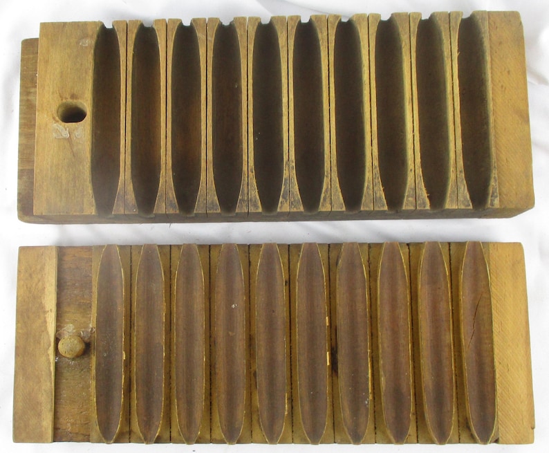 May include: A vintage wooden cigar mold consisting of two rectangular blocks with ten carved, elongated, concave channels. The top piece features a circular hole on the left side, while the bottom piece includes a small wooden peg for alignment.