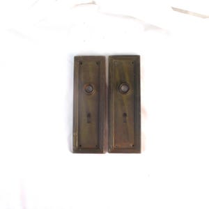 May include: Two rectangular brass door plates with a dark, aged patina. Each plate features a central circular opening for a door knob and a smaller keyhole opening below it. The plates have a stepped, linear border design around the edges.