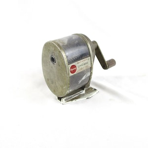 May include: Vintage silver-toned pencil sharpener with a wooden handle. The sharpener has a cylindrical body with a clear plastic window and a label that reads "Apso Chicago". The base is white.