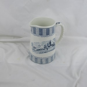 May include: A white ceramic pitcher with a blue and white patterned design. The pitcher features a scene of a village with a church and trees. The handle is twisted and has a blue and white pattern.