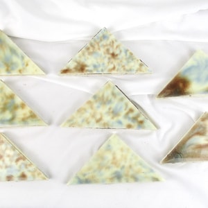 May include: Seven triangular ceramic tiles with a marbled pattern in shades of brown, blue, and cream. The tiles are arranged on a white fabric surface, showcasing their unique color variations and geometric shape.