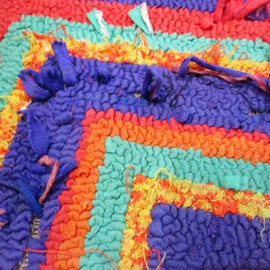 May include: Close-up of a colorful, multi-striped rug with a textured surface. The rug is made of woven fabric and has a frayed edge.