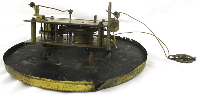 Peut inclure: Un mouvement d'horloge ancien avec des engrenages et des composants en laiton. Le m&eacute;canisme est mont&eacute; sur une base circulaire noire avec un bord dor&eacute;. Des fils et un pendule sont attach&eacute;s.