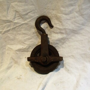 May include: A rusty metal pulley with a hook. The pulley has a single wheel with a groove for a rope. The hook is attached to the top of the pulley.