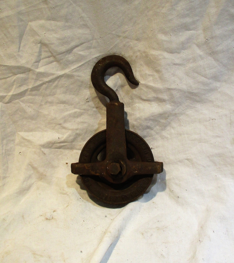 May include: A rusty metal pulley with a hook. The pulley has a single wheel with a groove for a rope. The hook is attached to the top of the pulley.