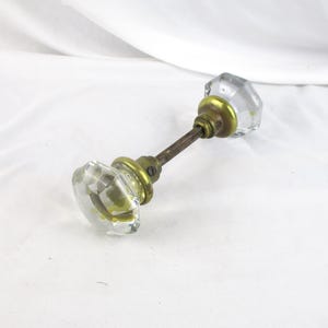 May include: A vintage door knob with clear, faceted glass knobs and brass hardware. The knobs are connected by a metal rod. The brass hardware shows signs of age. The door knob is a classic piece of hardware.