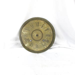 May include: A circular, weathered brass clock face featuring black Roman numerals arranged in a classic dial pattern. The center has a circular cutout, and two smaller circular holes are positioned near the bottom, showing signs of age and patina.
