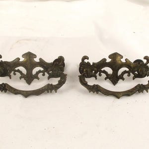 May include: Two antique brass drawer pulls with ornate floral designs. The pulls are in the shape of a half-circle with a decorative center piece.