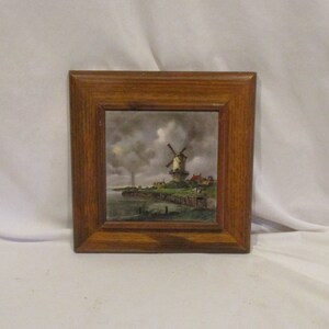 May include: A framed tile with a painting of a windmill in a Dutch landscape. The windmill is white with a red roof and is surrounded by green grass and a blue river. The sky is cloudy and gray.