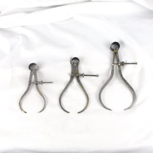 May include: Three antique metal calipers of varying sizes are arranged on a white background. Each caliper has curved legs, a central adjustment screw, and a rounded top. The calipers appear to be vintage tools.