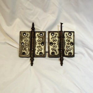 May include: A pair of antique, ornate, cast iron hinges with white paint and decorative details. The hinges are in good condition with some rust and wear.