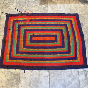 May include: A colorful woven rug with a concentric square design. The rug has a dark blue background with red, green, and yellow stripes.