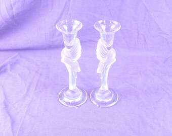 May include: Two clear glass candle holders with a frosted finish. The candle holders have a unique design with a swirling, sculpted shape. The base is round, and the top is a flared cup shape. The background is a solid lavender color.