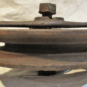 May include: A rusty metal pulley with two grooves, a metal frame, and a threaded bolt. The pulley is designed for use with rope or cable.