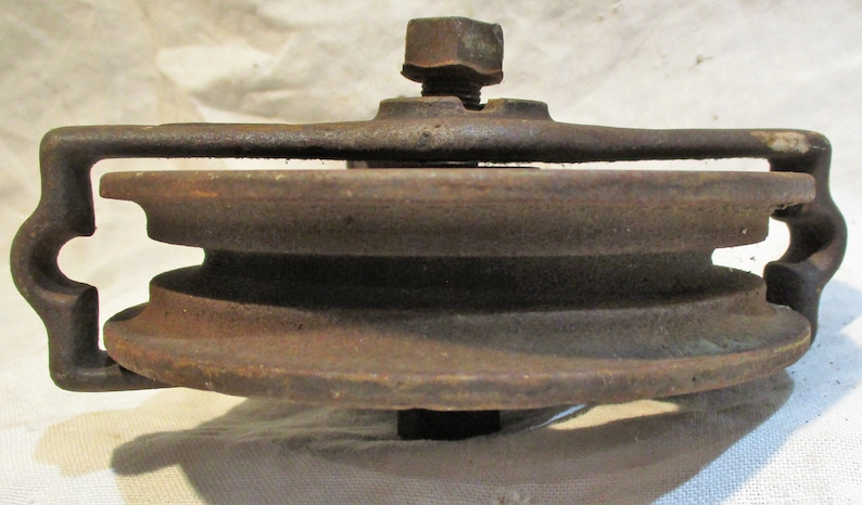 May include: A rusty metal pulley with two grooves, a metal frame, and a threaded bolt. The pulley is designed for use with rope or cable.