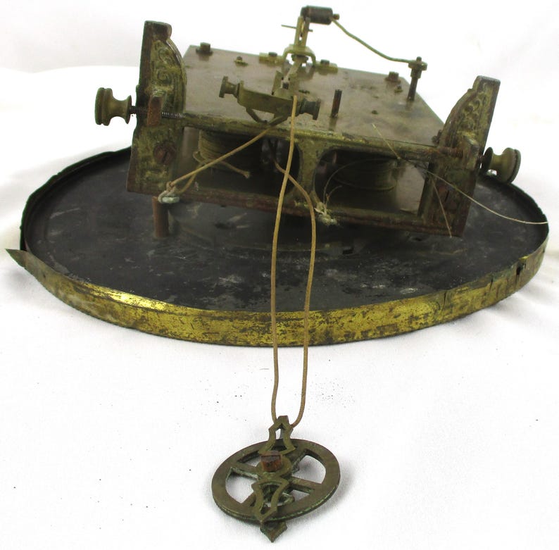 Peut inclure: M&eacute;canisme d'horloge antique en laiton et m&eacute;tal. Le m&eacute;canisme est mont&eacute; sur une base circulaire avec un bord dor&eacute;. Un pendule est suspendu &agrave; la face de l'horloge. Les composants de l'horloge montrent des signes d'&acirc;ge et d'usure.