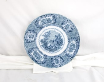 May include: A blue and white decorative plate with a scalloped edge. The plate features a central landscape scene and floral and architectural motifs around the rim. The plate is resting on a white cloth.