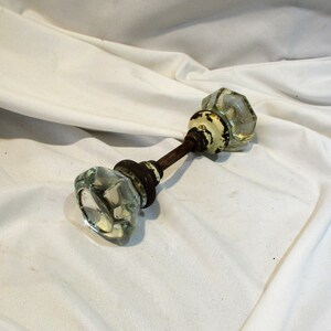 May include: Antique glass door knob with a metal rod connecting two knobs. The knobs are clear glass with a faceted design. The metal rod is rusty and has a white painted section.