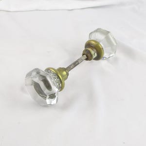 May include: A vintage clear glass door knob with a faceted design. The knob has a brass base and a tarnished metal rod connecting the two knobs. The door knob is set against a white background.