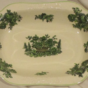 May include: A white oval serving dish with a green floral design. The design features a basket of flowers and leaves.
