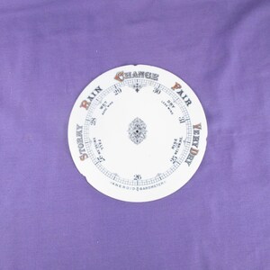 May include: A circular white aneroid barometer dial featuring black numerical markings from 26 to 31. The text includes labels such as STORMY, RAIN, CHANGE, FAIR, and VERY DRY, along with smaller text indicating weather conditions like WET and DRY.