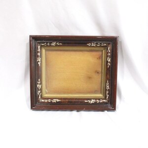 May include: A rectangular wooden picture frame with a dark brown finish and a polished sheen. The frame features inlaid white floral patterns along its border and a gold-toned inner rim surrounding a plain, light tan wooden center panel.