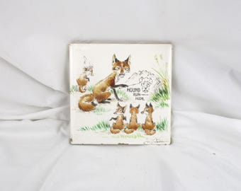 May include: A vintage ceramic tile with a hand-painted illustration of a fox family. The mother fox is teaching her kits to "hound run hide". The tile is white with a green and brown background.
