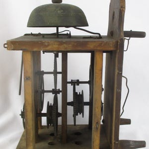 May include: Antique wooden clock mechanism with visible gears and a bell. The clock has a rectangular wooden frame with a dark metal bell on top. The gears are visible inside the frame. The clock is made of wood and metal.