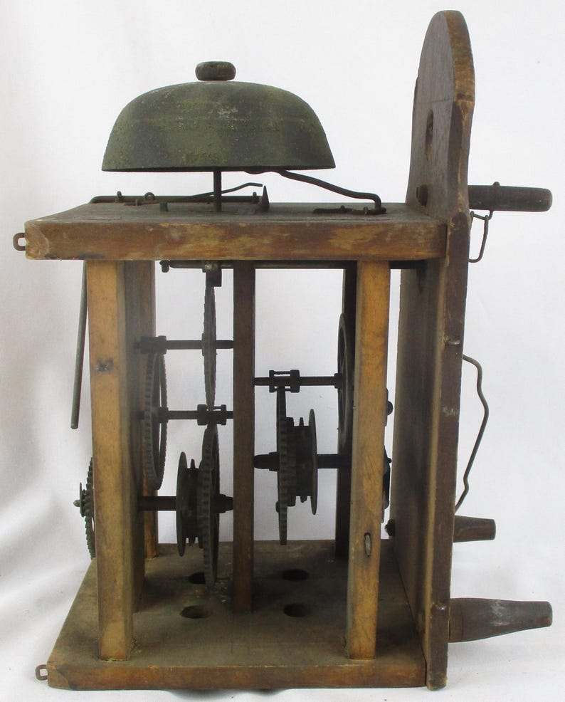 May include: Antique wooden clock mechanism with visible gears and a bell. The clock has a rectangular wooden frame with a dark metal bell on top. The gears are visible inside the frame. The clock is made of wood and metal.