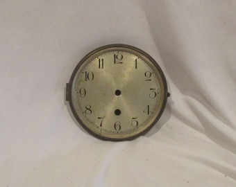 May include: A round, silver clock face with black Roman numeral markings for the hours. The clock face has two holes in the centre.
