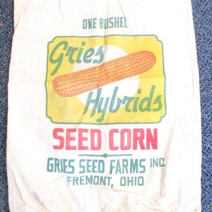 May include: A white canvas bag with the words "ONE BUSHEL" at the top. The bag features a yellow and green graphic with the words "Gries Hybrids" and an illustration of an ear of corn. Below, the text "SEED CORN" is in red, and "GRIES SEED FARMS INC. FREMONT, OHIO" is in green.