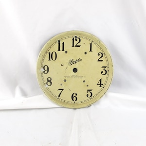 May include: A vintage clock face with black numerals on a cream-colored background. The word "Simplex" is printed in cursive, and the text "Simplex Time Recorder Co." is also visible. The clock face is round.