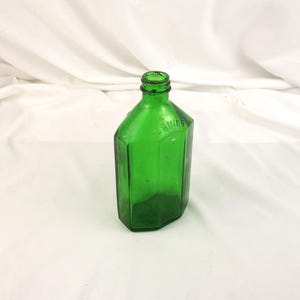 May include: A vintage, emerald green glass bottle with a hexagonal shape. The bottle has a narrow neck and a slightly raised shoulder. The word "SQUIBB" is embossed on the bottle. The bottle is empty and sits on a white surface.